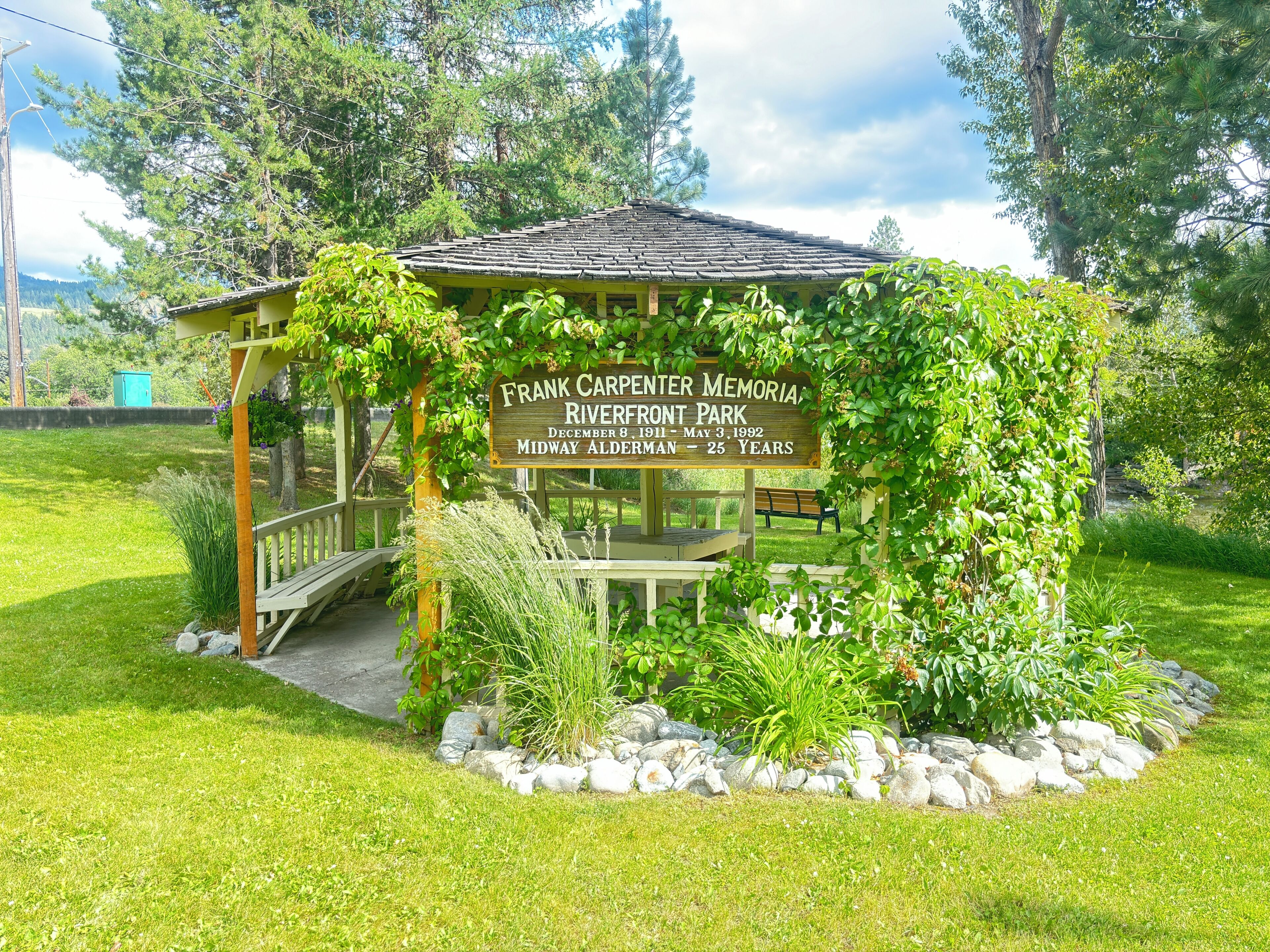 A memorial gazebo with a plaque, surrounded by greenery and a stone border.