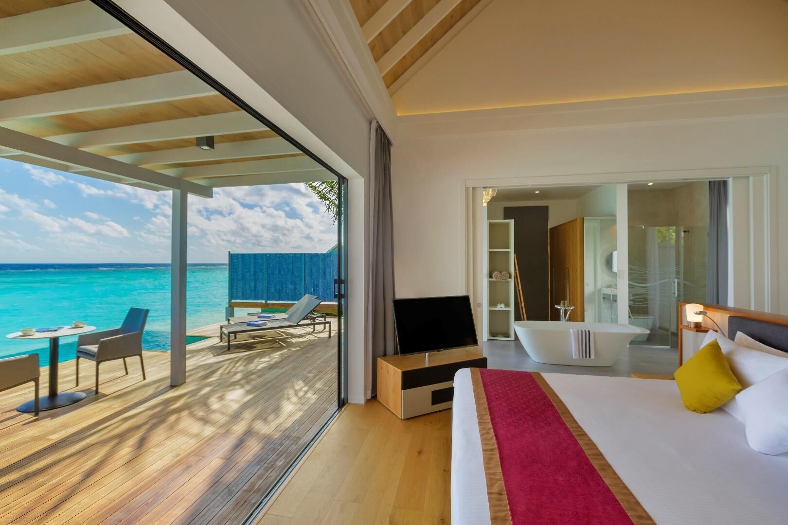 A modern hotel room with a large bed, a TV, a bathroom with a bathtub, and a balcony with a view of the ocean.