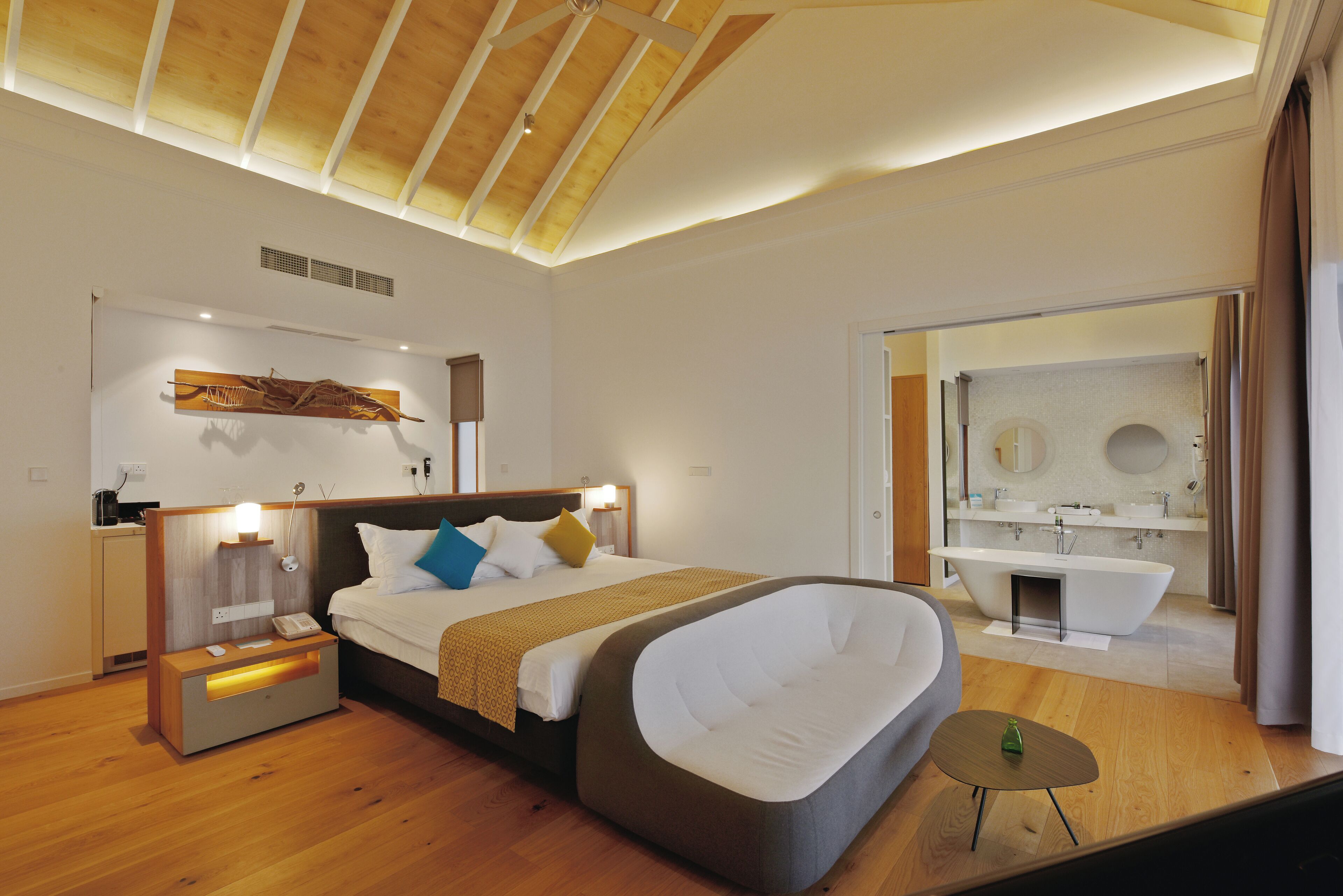 A modern hotel room with a large bed, a built-in bathtub, a separate bathroom with a large mirror, and a wooden ceiling.