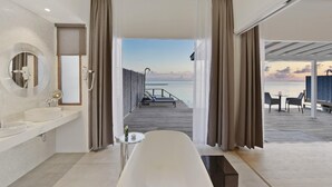 A modern bathroom with a bathtub, a round mirror, and a sink. A wooden deck leads to a balcony with a view of the sea. - Niva Kuramathi Maldives (Kuramathi Island)