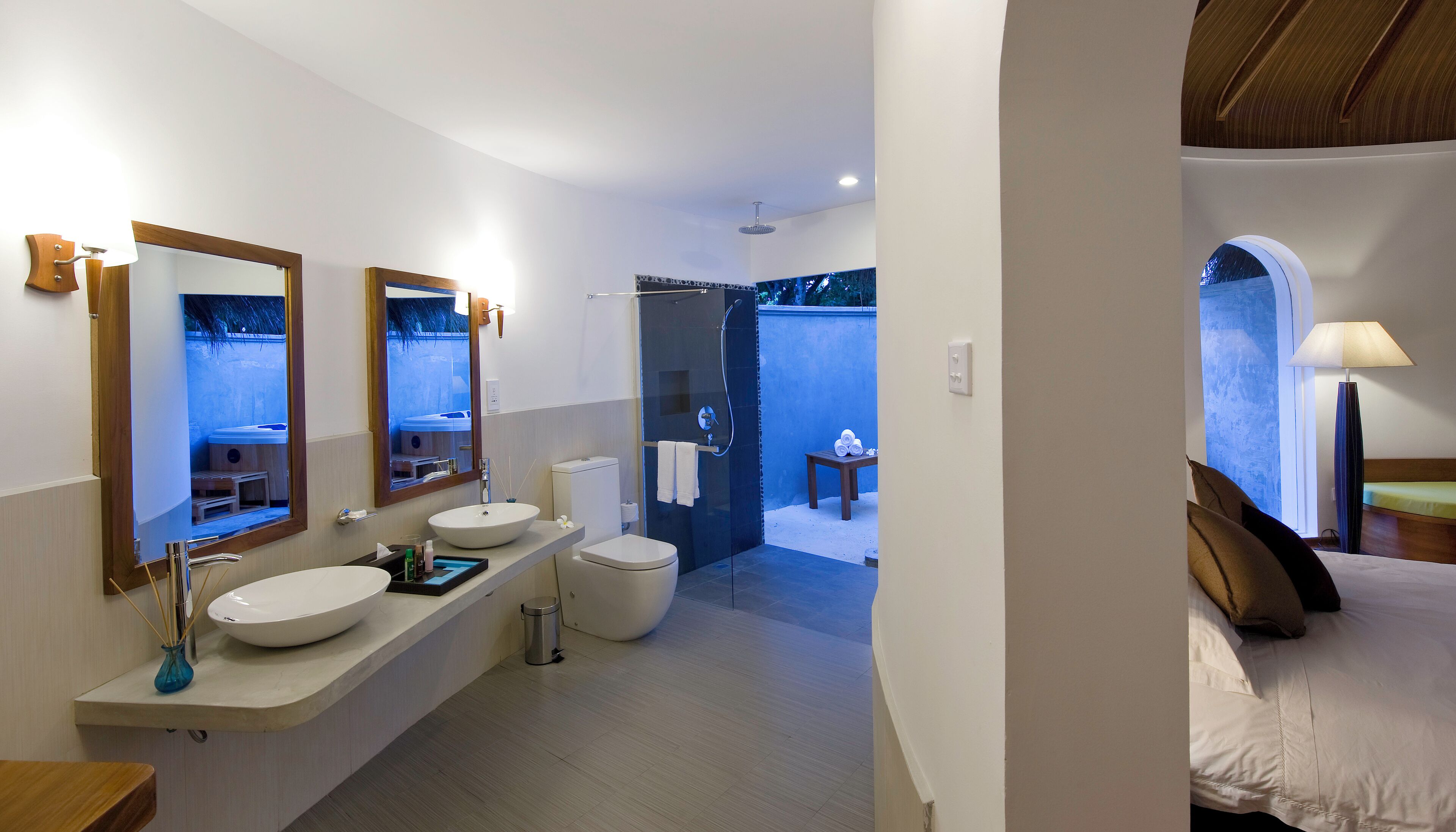 A modern bathroom with two sinks, a shower, and a toilet. There is a small table with towels and a trash can.