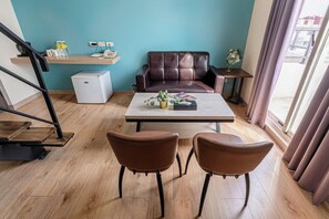 Een moderne woonkamer met een bruine leren bank, een salontafel en twee fauteuils. Tegen de muur staat een klein wit kastje, en er is een raam met gordijnen zichtbaar.