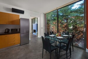 Una cocina moderna con refrigerador, microondas y gabinetes de madera. Hay un comedor con una mesa de vidrio y sillas, y un ventanal con vista a árboles.