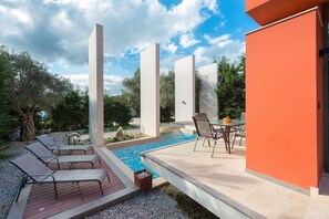 Un área de piscina moderna al aire libre con sillones de descanso, una mesa con fruta y un cielo despejado.