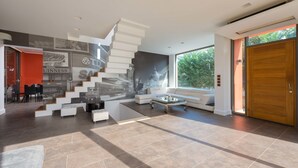 A spacious living area with a modern staircase, a seating area with a glass coffee table, and a large window offering a view of greenery. - Almond Villa (Rhodes)