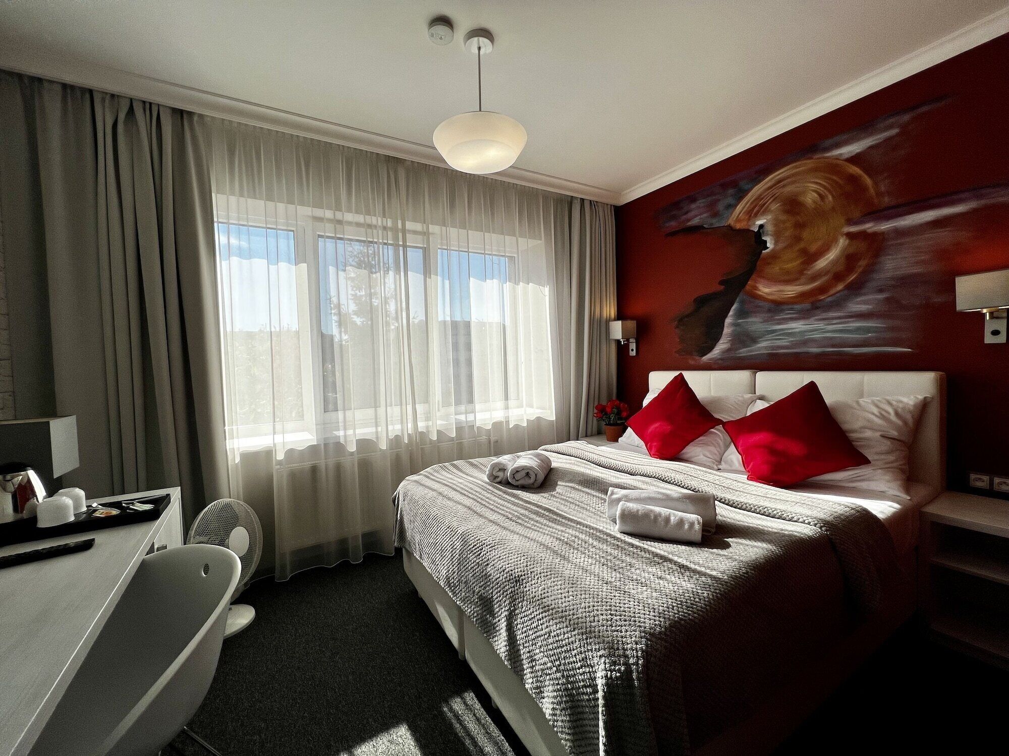A hotel room with a bed, a desk, a chair, a window with curtains, a painting on the wall, and a ceiling light.
