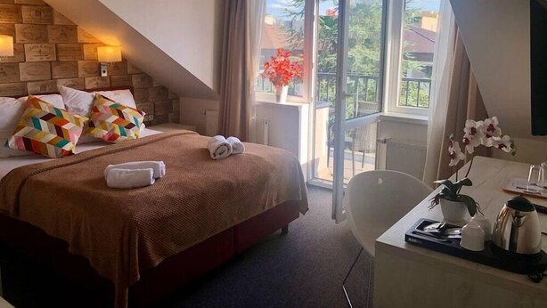 A hotel room with a bed, a desk with a plant, a window with curtains, and a balcony with a view of trees and buildings. - Hotel Shato (Prague)