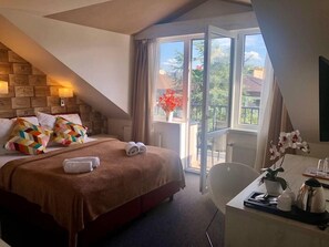 Habitación de hotel con cama, escritorio con planta, ventana con cortinas y balcón con vista a árboles y edificios.