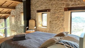A bedroom with a stone wall, a wooden ceiling, a bed with a grey bedspread, and a chair. - Le Pailler - DIO-ET-VALQUIERES (DIO-ET-VALQUIERES)
