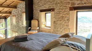 A bedroom with a stone wall, wooden ceiling beams, a bed with pillows, and a chair. - Le Pallier + extension (DIO-ET-VALQUIERES)