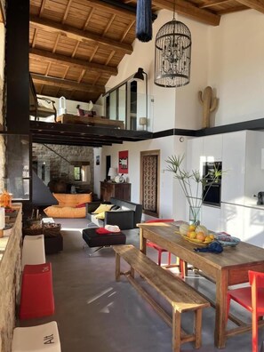 A spacious living area with a wooden ceiling, a dining table with chairs, a sofa, and a kitchen area in the background. - Le Pallier + extension (DIO-ET-VALQUIERES)