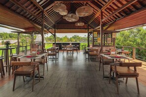 Un restaurant a l'aire lliure amb taules i cadires de fusta, un sostre alt amb lluminàries teixides i vistes a la vegetació.