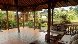 A covered patio with a wooden bench and a table, surrounded by lush greenery and a view of a traditional house. - Hideaway Villas Candy Ubud (Ubud)