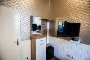 Habitación pequeña con una puerta blanca, un mueble de madera con cajones, un espejo y un televisor de pantalla plana montado en la pared.