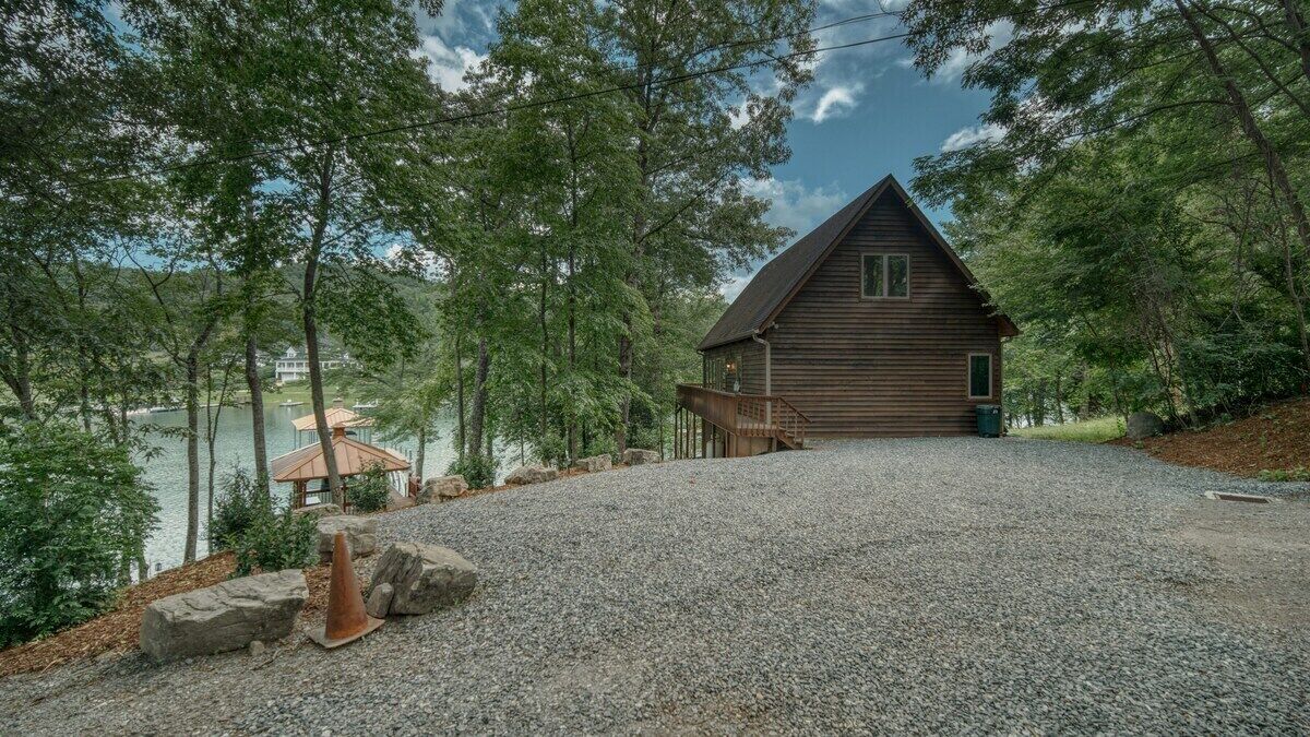 A cabin with a gravel driveway, surrounded by trees and a lake.