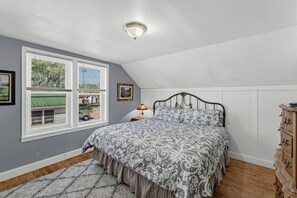 Une chambre à coucher comprenant un lit, une commode, une fenêtre avec des stores, une lampe et un luminaire au plafond.