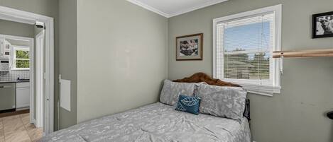 Un dormitorio con cama, una ventana con persianas, un cuadro en la pared y una cocina visible a través de una puerta abierta.