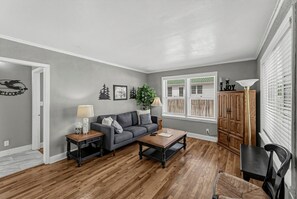Un salon avec un canapé gris, une table basse en bois et une lampe. On y trouve une fenêtre avec des stores, un meuble et une plante.