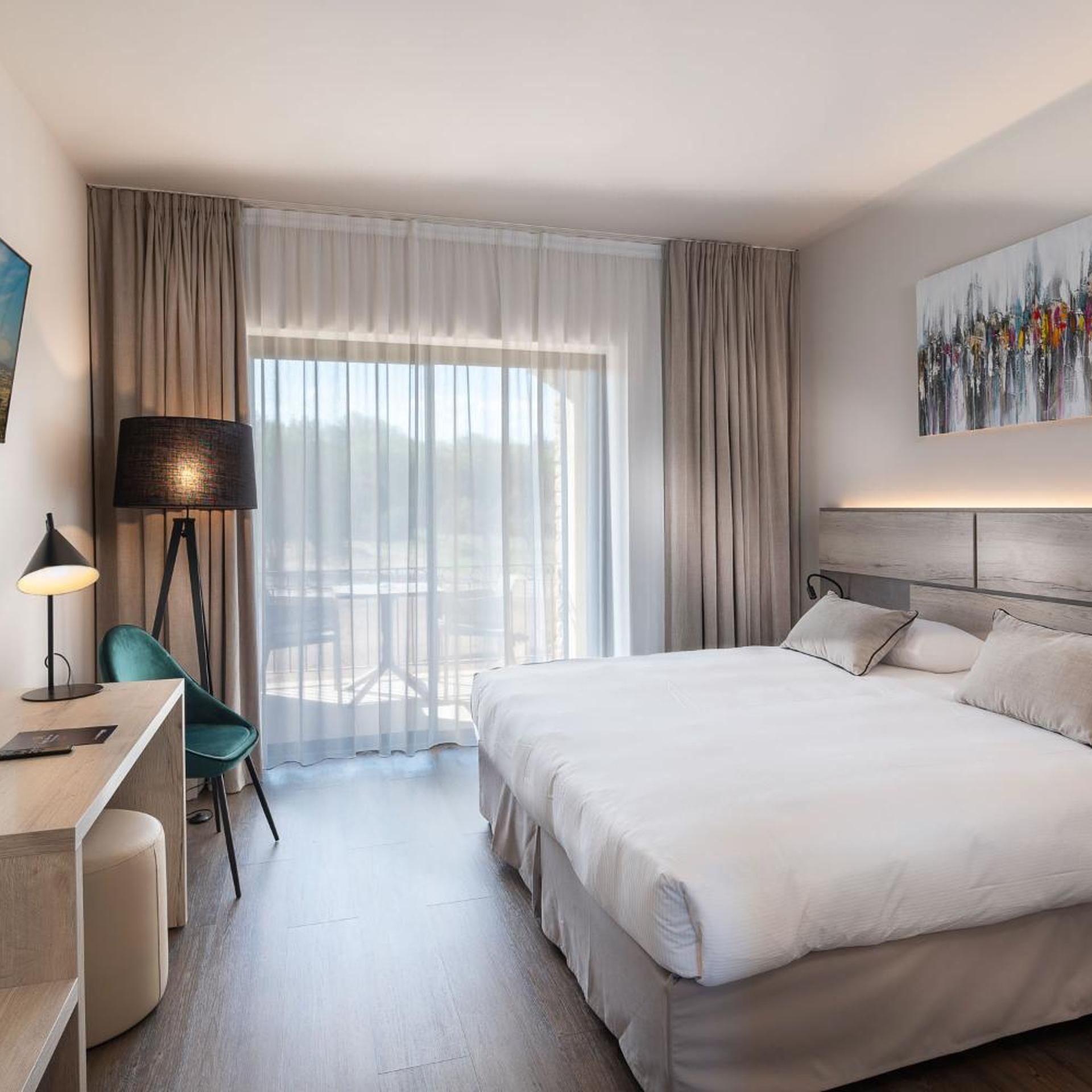 A hotel room with a bed, desk, chair, and a painting on the wall.