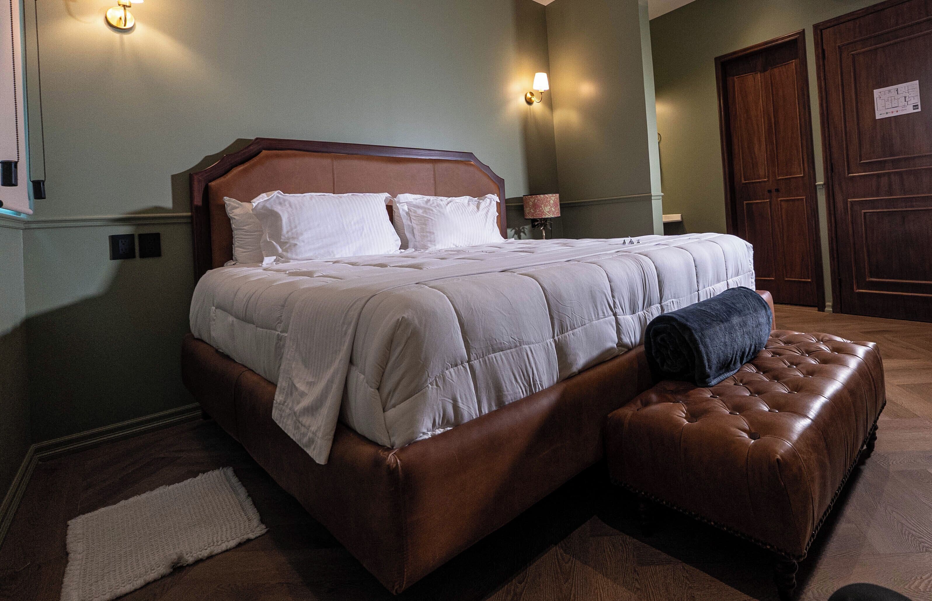 Una cama bien hecha con cabecera, lámpara de noche y un reposapiés de cuero marrón.