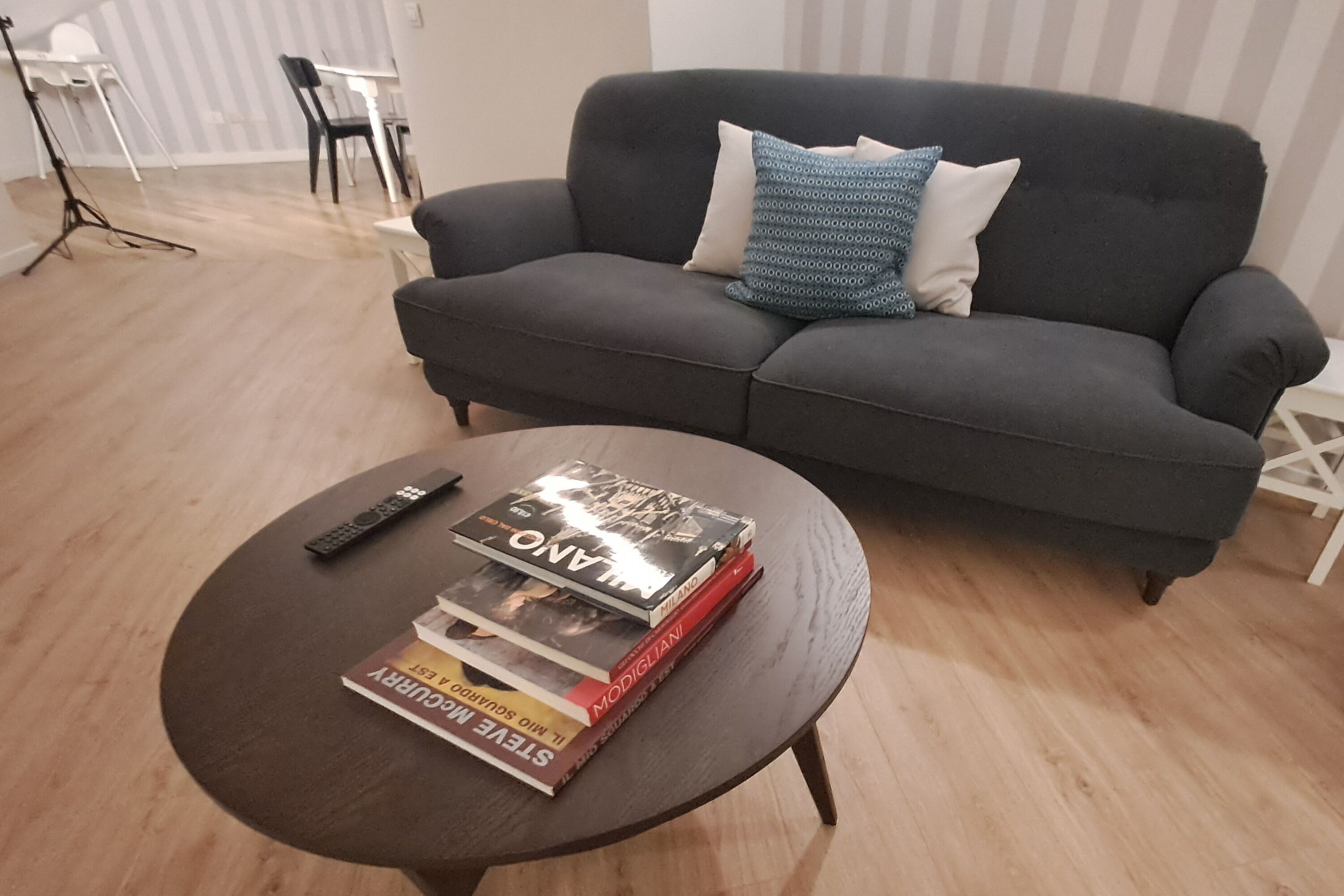 Ein Wohnzimmer mit einem dunkelgrauen Sofa, einem runden Couchtisch mit Büchern und einer Fernbedienung, sowie einer gestreiften Wand im Hintergrund.