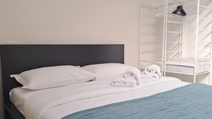 Ein ordentlich gemachtes Bett mit weißen Leinen und einer blauen Decke, ein weißes Handtuch liegt gefaltet auf dem Bett, und ein weißes Regal mit einem aufgehängten Gegenstand.