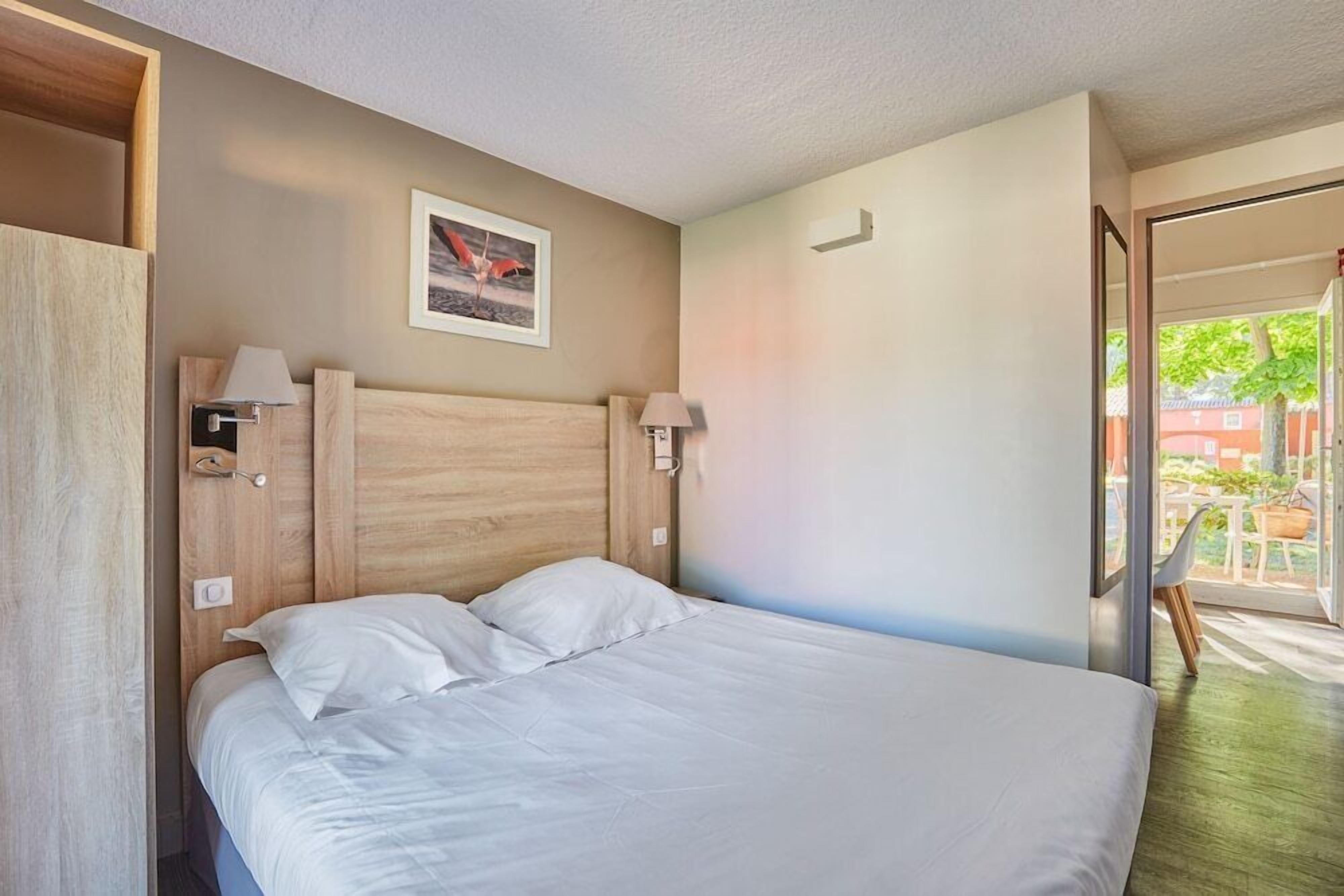 Ein Hotelzimmer mit einem Holzkopfteil, zwei Nachttischlampen, einem gerahmten Bild an der Wand und Blick ins Freie durch eine Glastür.