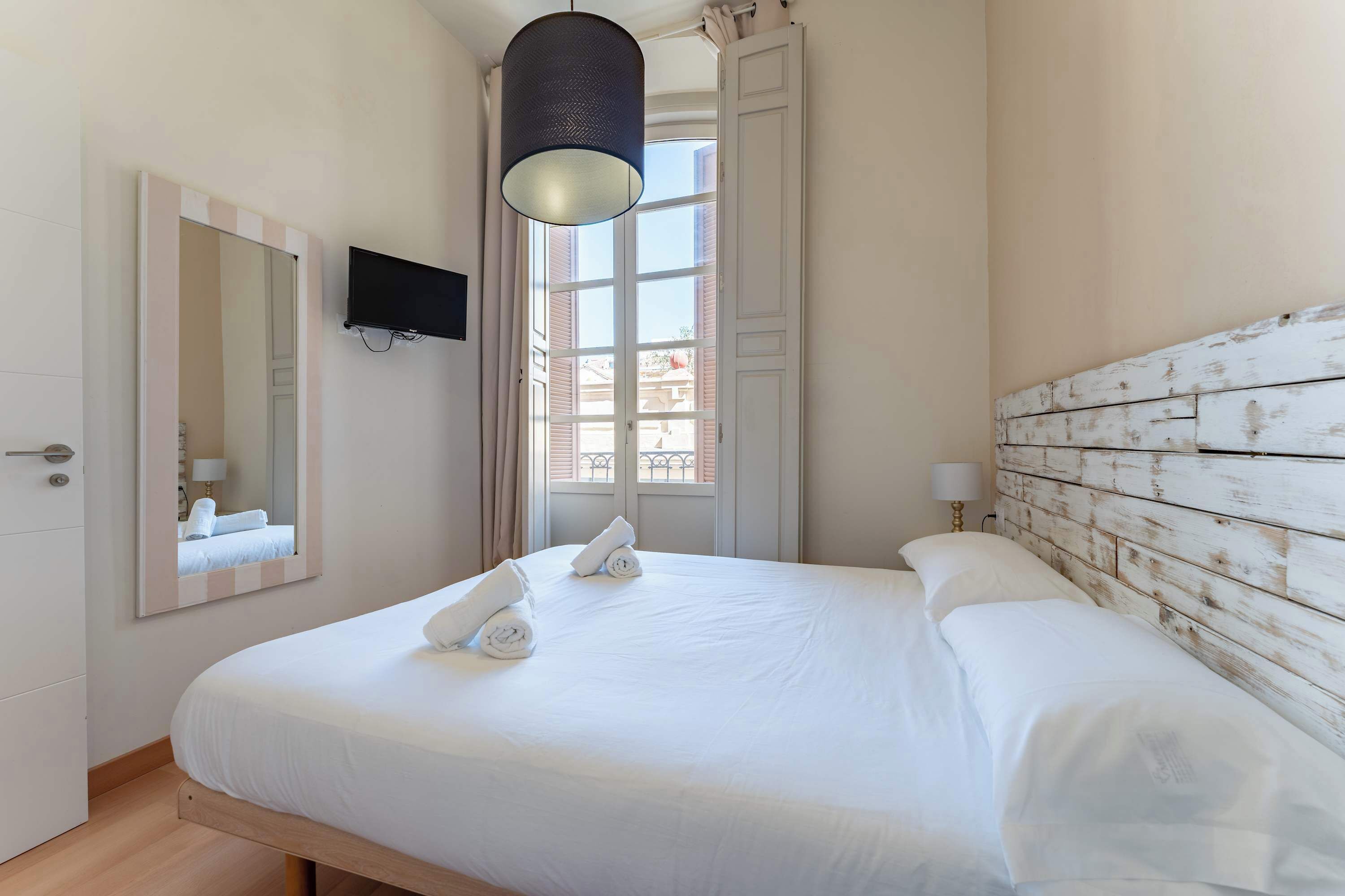 A neatly made bed with white linens and towels, a television mounted on the wall, and a window with curtains.