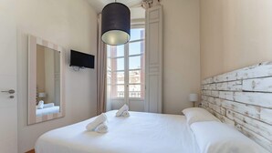 Ein ordentlich bezogenes Bett mit weißen Leinen und Handtüchern, ein an der Wand befestigter Fernseher und ein Fenster mit Vorhängen.