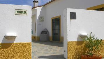 A white building with a yellow base, an entrance sign, a door, and a small garden area with a potted plant.