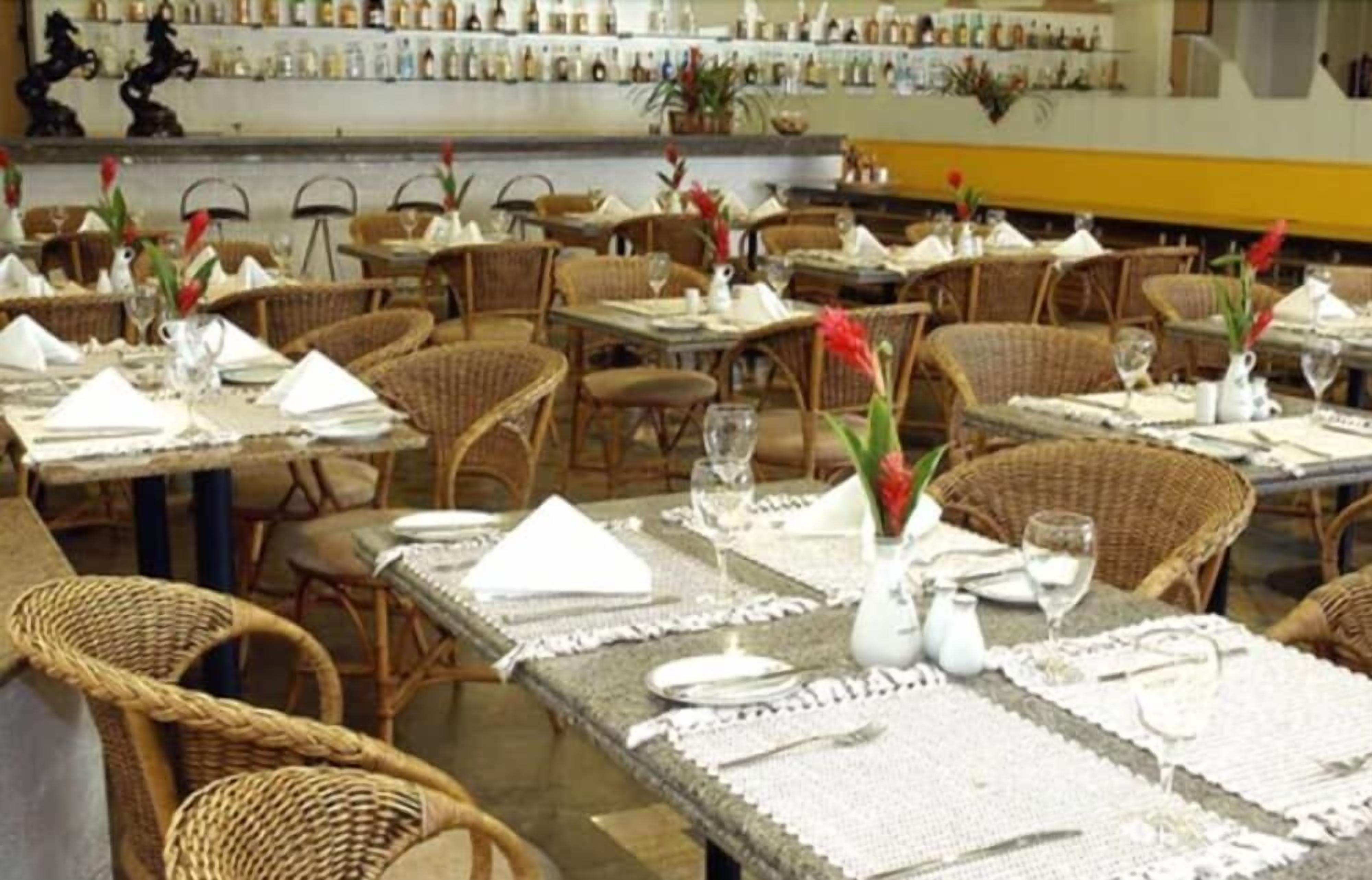 Restaurante com mobiliário de vime, mesas bem arrumadas com guardanapos brancos e um bar ao fundo.
