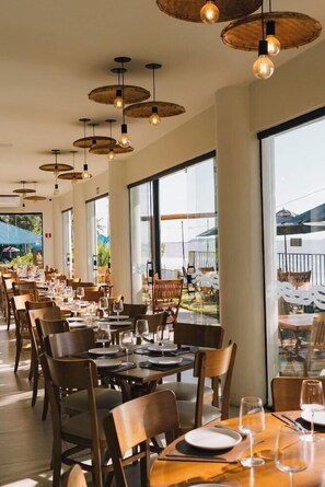 A restaurant with multiple tables and chairs, large windows, and hanging light fixtures.