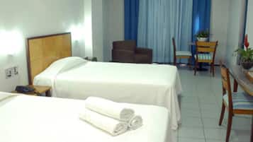 Quarto de hotel com duas camas, uma cadeira, uma mesinha e um vaso de planta.