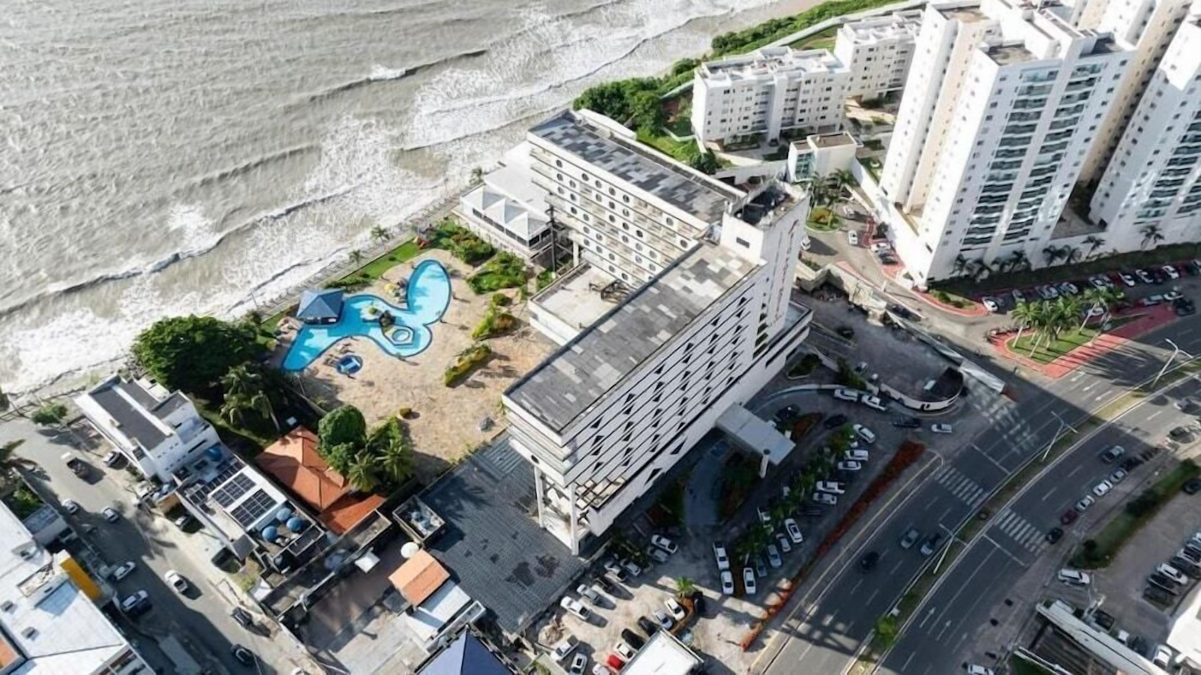 Vista aérea de um hotel à beira-mar, com piscina, estacionamento e edifícios altos adjacentes.