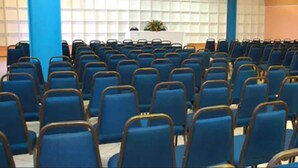 Une salle de conférence avec des rangées de chaises bleues, une table blanche ornée d’une plante et des unités de climatisation fixées aux murs.