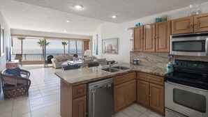 A modern kitchen with a dishwasher, double sink, and stainless steel appliances. It features wooden cabinets and a large window overlooking a balcony with a view of the ocean. - Oceanfront Sunsets at Sonoran Spa E305 (Puerto Peñasco)