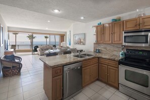 A modern kitchen with a dishwasher, double sink, and stainless steel appliances. It features wooden cabinets and a large window overlooking a balcony with a view of the ocean. - Oceanfront Sunsets at Sonoran Spa E305 (Puerto Peñasco)
