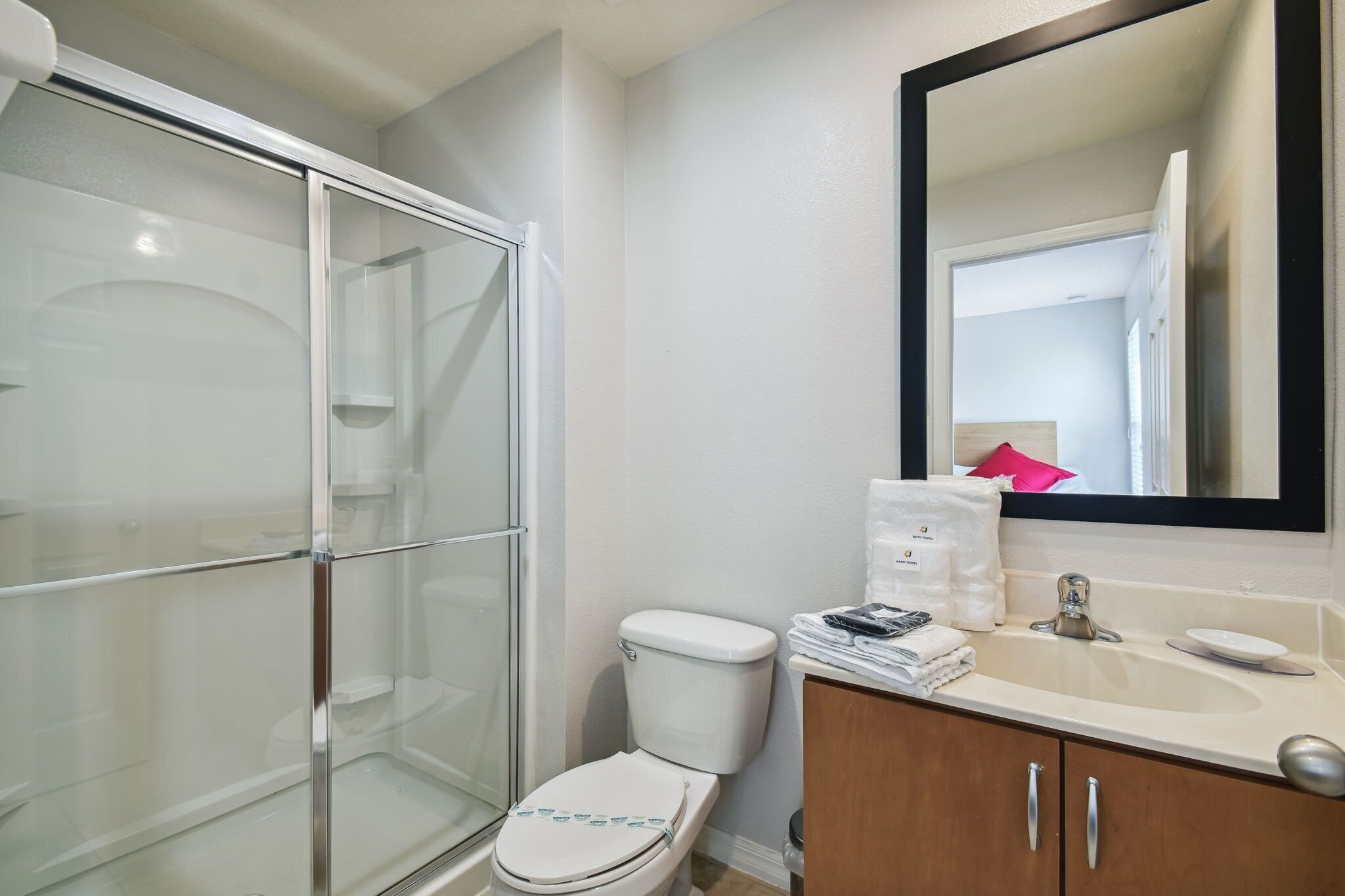 A bathroom with a glass shower enclosure, a toilet, a sink with a mirror above it, and a cabinet with toiletries.
