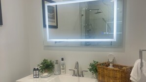 A bathroom with a white vanity, a large mirror with a light, two sinks, wooden drawers, a wicker basket, and a towel rack. - Cozy Private Room 2 - Your Quiet Downtown Miami Getaway (Miami)