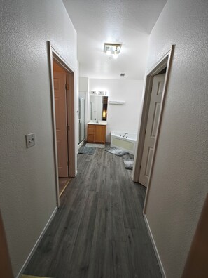 Un pasillo con piso de madera, paredes blancas y un baño con lavamanos y espejo.