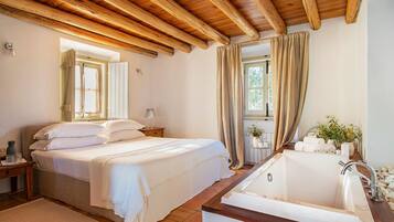 A bedroom with a large bed, a wooden ceiling, a window with curtains, and a bathroom with a sink and bathtub.