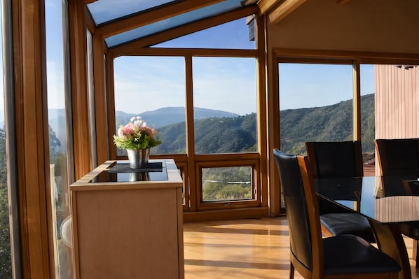 A modern room with large windows offering a mountain view, a wooden desk with a vase of flowers, and a dining area with chairs.