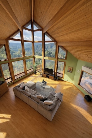 A spacious living room with a large sectional sofa, a flat-screen TV, and a wooden ceiling with large windows offering a view of the mountains.