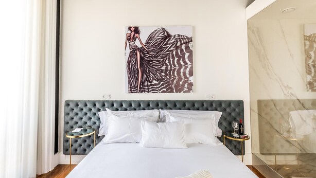 A neatly made bed with white linens, a grey tufted headboard, and a single white pillow. There is a small round table with a gold base and a glass top, holding a bottle and glasses. A large abstract painting hangs on the wall above the bed. - RM Guest House The Experience (Setubal)
