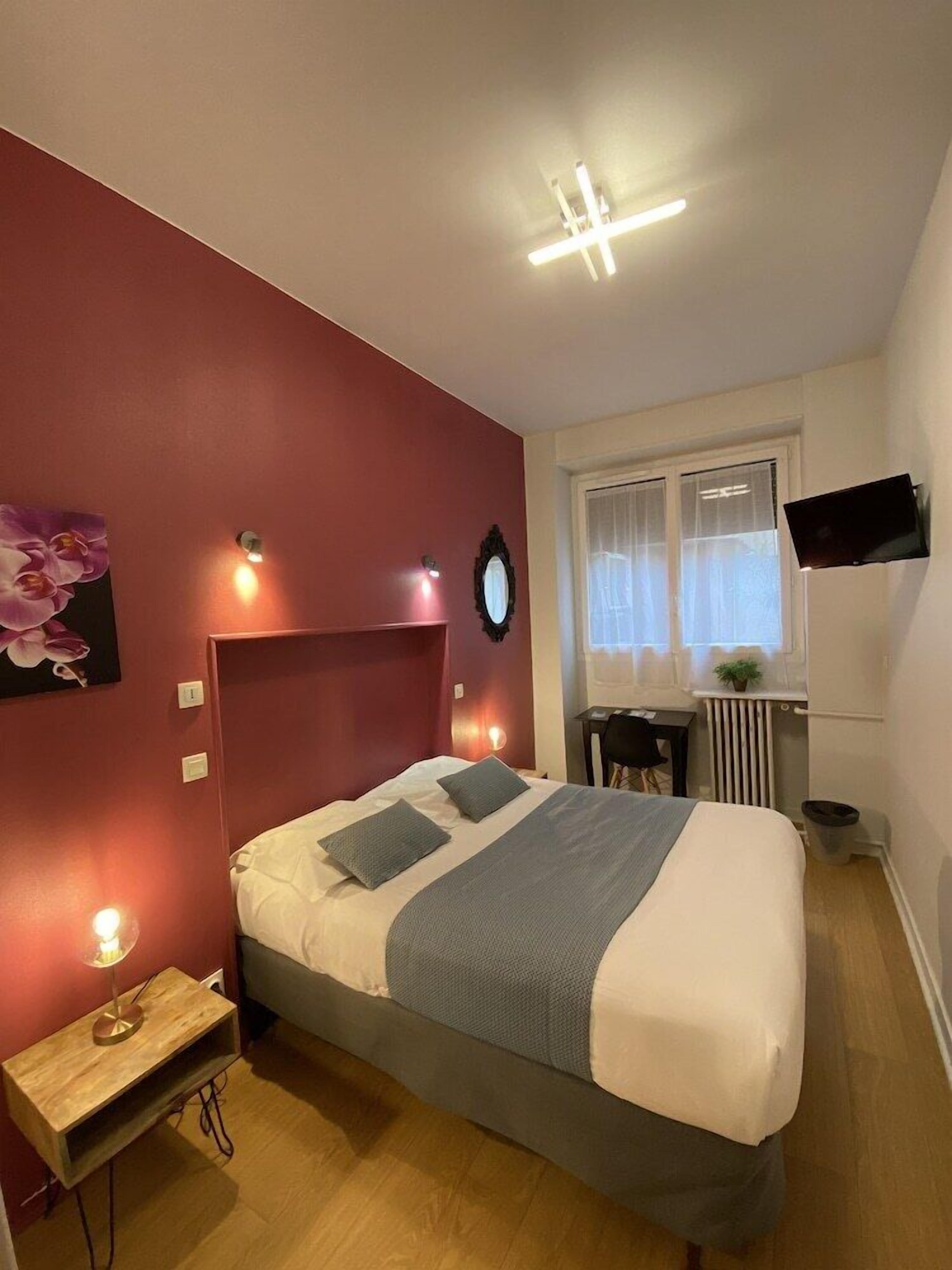 A bedroom with a bed, bedside table, lamp, wall-mounted TV, and a window with curtains.