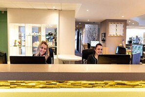 A reception area with two receptionists at computers, a glass display case with items, and a wall with a bulletin board. - Domaine de La Foret D Orient Natur Hotel Golf Spa (Rouilly-Sacey)