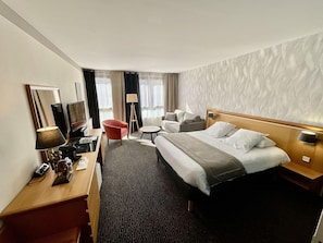 A hotel room with a large bed, a desk with a lamp, a TV, a red chair, and a sofa. - Domaine de La Foret D Orient Natur Hotel Golf Spa (Rouilly-Sacey)