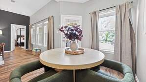 Un espace repas moderne avec une table ronde, des chaises recouvertes d’un tissu vert, un vase orné de fleurs violettes et une vue sur l’extérieur grâce à de grandes fenêtres.