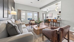Un vaste salon comprenant un canapé, une table basse et un fauteuil en cuir. On y trouve également un coin repas avec une table et des chaises, ainsi qu’une cuisine à l’arrière-plan.