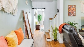 A hallway with a white sofa, colorful cushions, a macrame wall hanging, a staircase, and a potted plant.